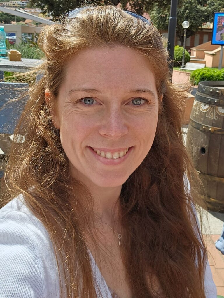 Lächelnde Coachin Mandy mit natürlichen roten Haaren im Sonnenschein – einladend und inspirierend, Coaching für kreative Journals und Körperarbeit.